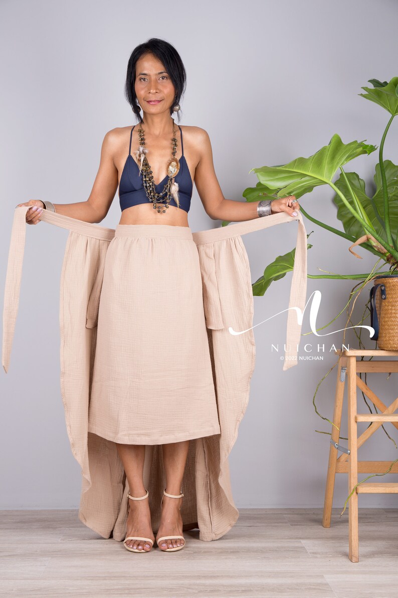 May include: A woman wearing a beige wrap skirt with a long train, a navy blue bra, and a layered necklace. The skirt is tied at the waist and has a high-low hemline. The woman is standing in front of a grey background with a potted plant to the right.