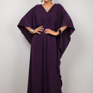 May include: A woman wearing a long, purple kaftan dress with a gathered neckline and flowing sleeves. She is standing with her hands on her hips and is wearing a silver bracelet and a silver necklace.