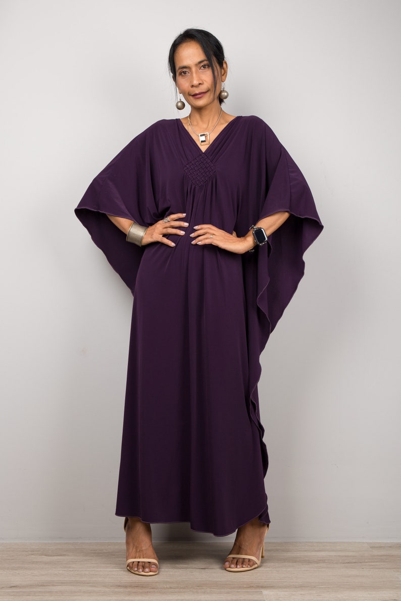May include: A woman wearing a long, purple kaftan dress with a gathered neckline and flowing sleeves. She is standing with her hands on her hips and is wearing a silver bracelet and a silver necklace.