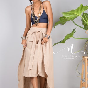May include: A woman wearing a navy blue bra top with a layered beaded necklace and a long, beige wrap skirt. The skirt has a high-low hemline and is tied at the waist with a sash. The woman is standing in front of a grey wall with a green plant in the background.