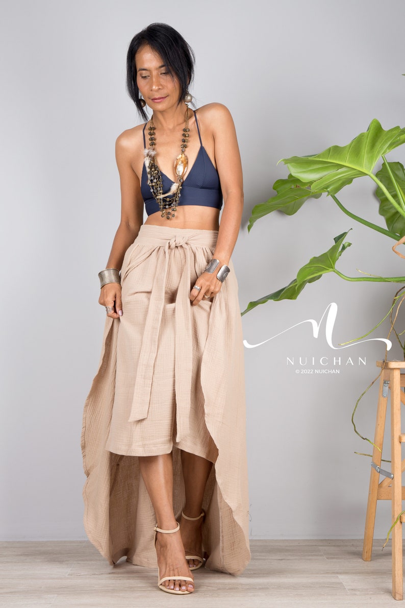 May include: A woman wearing a navy blue bra top with a layered beaded necklace and a long, beige wrap skirt. The skirt has a high-low hemline and is tied at the waist with a sash. The woman is standing in front of a grey wall with a green plant in the background.