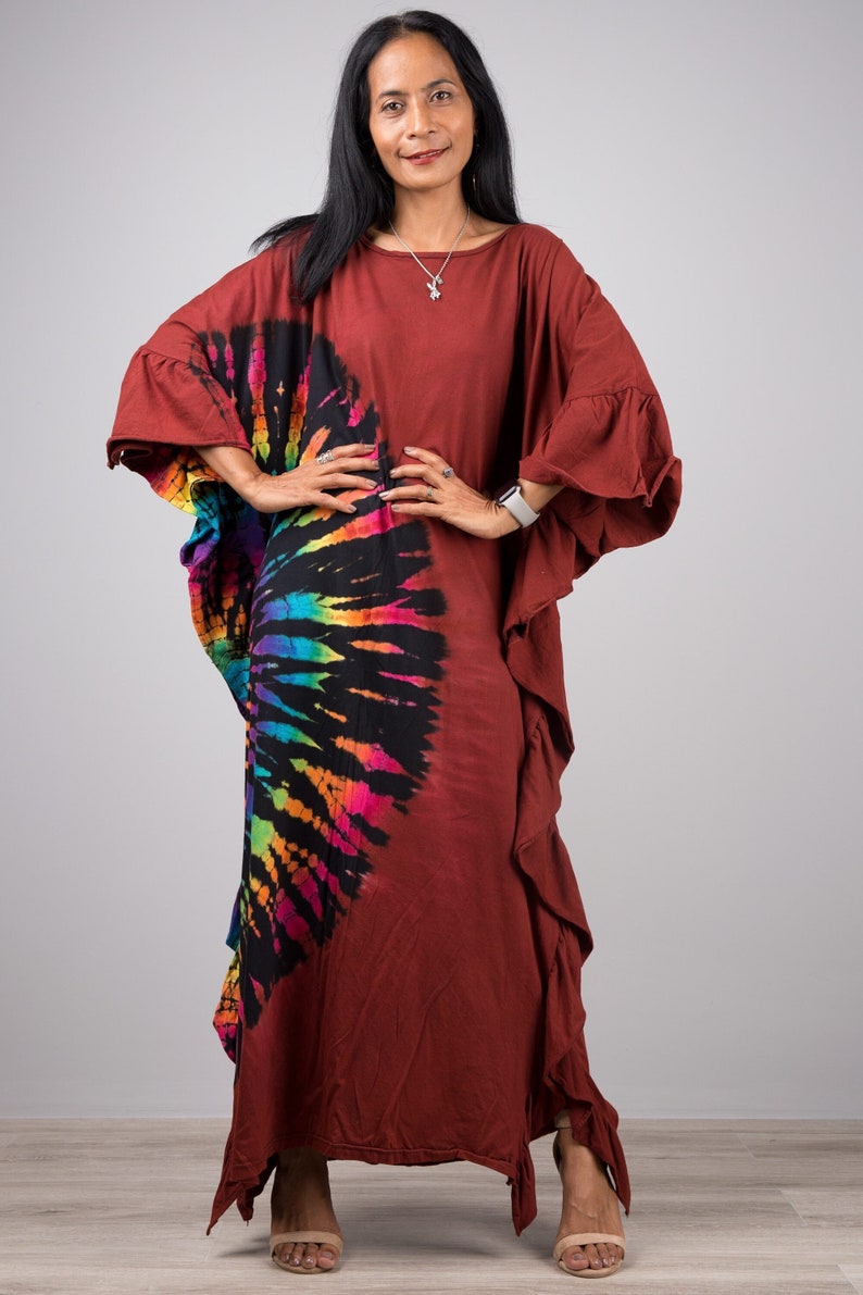May include: A long, maroon kaftan with a rainbow tie-dye design on the left side. The kaftan has a ruffled hem and a relaxed fit. The model is wearing the kaftan with a silver necklace and a watch.