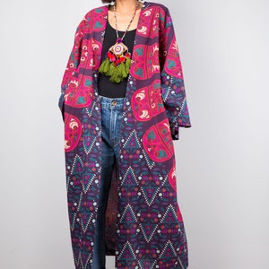 Pu&ograve; includere: Un lungo kimono con motivo viola e rosa con una collana di nappe verdi. Il kimono ha un motivo geometrico con un design floreale. La modella indossa jeans blu e stivali neri.