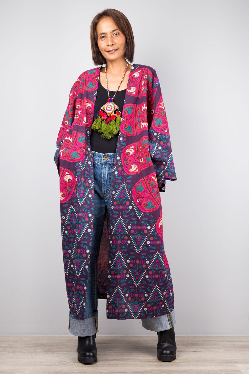 Pu&ograve; includere: Un lungo kimono con motivo viola e rosa con una collana di nappe verdi. Il kimono ha un motivo geometrico con un design floreale. La modella indossa jeans blu e stivali neri.