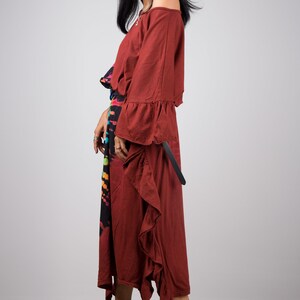 May include: A woman wearing a maroon off-the-shoulder dress with a ruffled hem and long sleeves. She is wearing a black and rainbow tie-dye skirt underneath the dress. She is wearing brown heeled sandals.