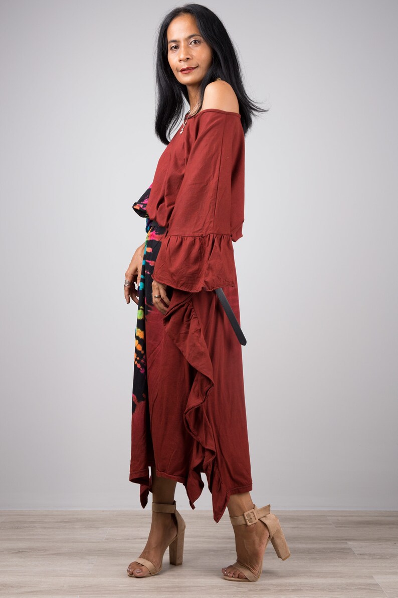May include: A woman wearing a maroon off-the-shoulder dress with a ruffled hem and long sleeves. She is wearing a black and rainbow tie-dye skirt underneath the dress. She is wearing brown heeled sandals.