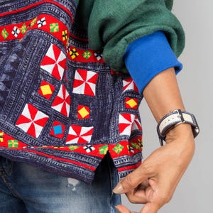 K&ouml;nnte beinhalten: Ein gr&uuml;n-blaues gemustertes Hemd mit roten, wei&szlig;en und gelben Stickereien. Das Hemd hat einen blauen &Auml;rmelabschluss und wird &uuml;ber einer blauen Jeans getragen.