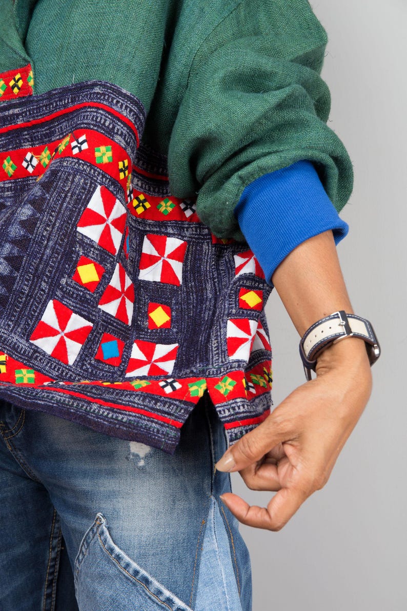 K&ouml;nnte beinhalten: Ein gr&uuml;n-blaues gemustertes Hemd mit roten, wei&szlig;en und gelben Stickereien. Das Hemd hat einen blauen &Auml;rmelabschluss und wird &uuml;ber einer blauen Jeans getragen.