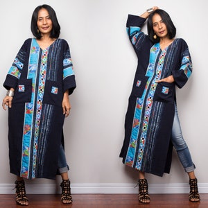 以下が含まれることがあります： Vネックのカラフルな幾何学模様が施された、長いダークブルーのチュニック。チュニックは長袖で、大きなポケットが2つ付いています。モデルはチュニックにジーンズと黒いサンダルを合わせています。