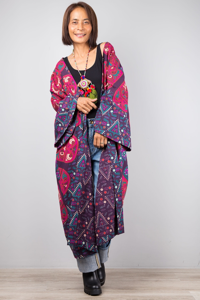 Pu&ograve; includere: Una donna indossa un kimono lungo con motivi viola e rosa, un top nero e jeans blu. Indossa una collana di perline colorate con un fiocco.