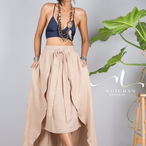 May include: A woman wearing a beige wrap skirt with a high-low hemline, a navy blue halter top, and a layered necklace with feathers and beads. The skirt has a tie at the waist and is made of a soft, flowing fabric. The woman is standing in front of a grey wall with a plant in the background.