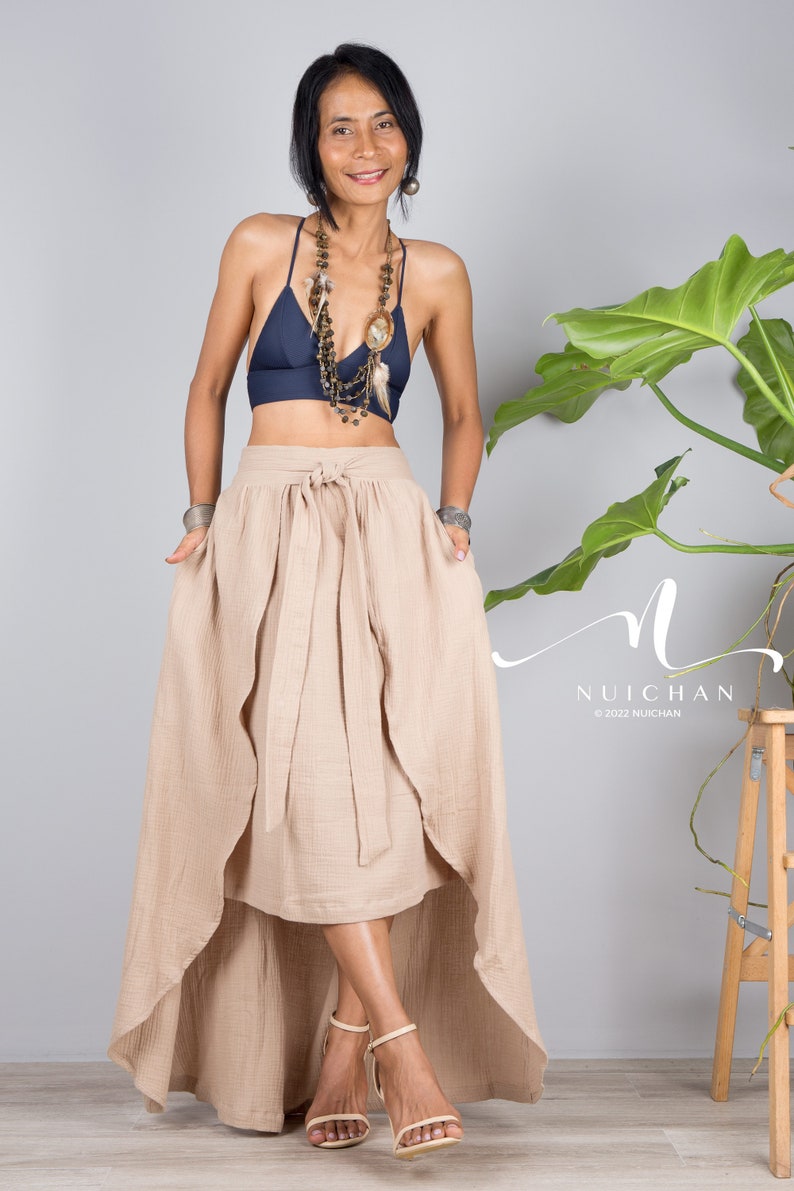 May include: A woman wearing a beige wrap skirt with a high-low hemline, a navy blue halter top, and a layered necklace with feathers and beads. The skirt has a tie at the waist and is made of a soft, flowing fabric. The woman is standing in front of a grey wall with a plant in the background.