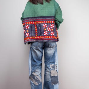 K&ouml;nnte beinhalten: Eine gr&uuml;ne und blaue gemusterte Jacke mit roten Stickereien. Die Jacke wird &uuml;ber einer blauen Jeans mit Patches getragen.