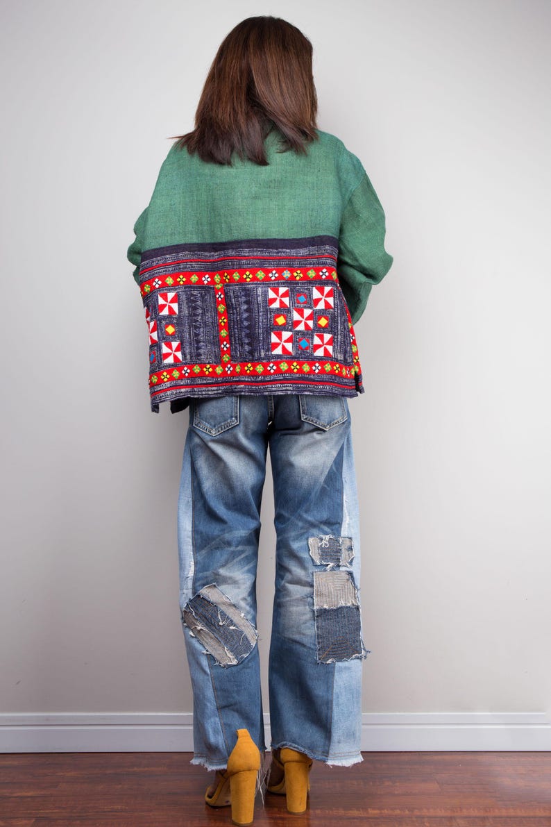 K&ouml;nnte beinhalten: Eine gr&uuml;ne und blaue gemusterte Jacke mit roten Stickereien. Die Jacke wird &uuml;ber einer blauen Jeans mit Patches getragen.