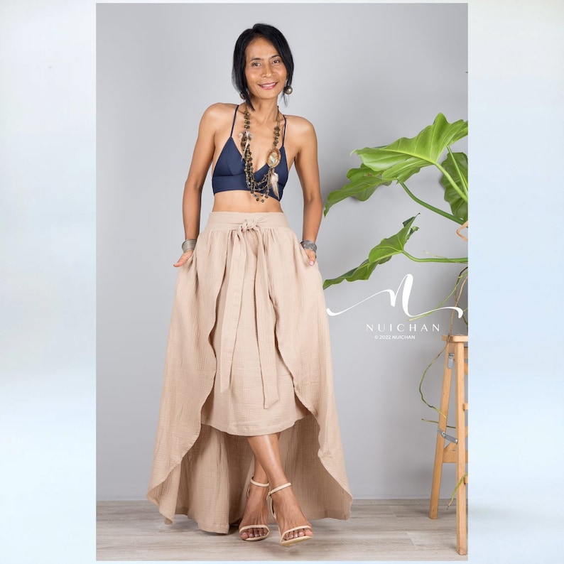 May include: A woman wearing a beige high-low skirt with a tie at the waist, a navy blue crop top, and a long beaded necklace. The skirt has a flowing, layered design.