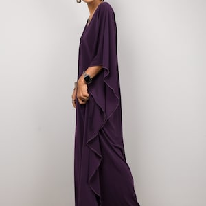 May include: A woman wearing a long, flowing purple kaftan with a ruffled hem. She is wearing a watch on her wrist and has her hair pulled back.