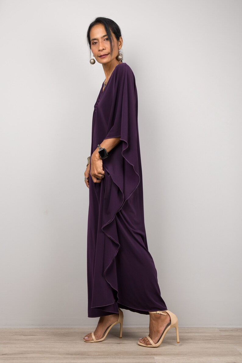 May include: A woman wearing a long, flowing purple kaftan with a ruffled hem. She is wearing a watch on her wrist and has her hair pulled back.