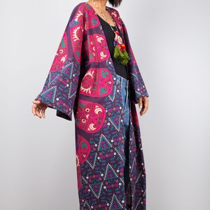 Pu&ograve; includere: Un kimono lungo e fluido di colore viola e rosa con un motivo geometrico. Il kimono ha maniche lunghe ed &egrave; aperto sul davanti. La modella indossa il kimono sopra una maglietta nera e jeans blu.
