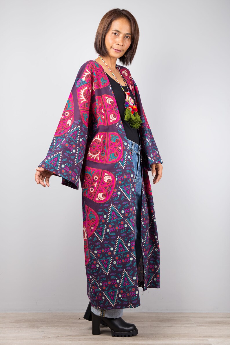 Pu&ograve; includere: Un kimono lungo e fluido di colore viola e rosa con un motivo geometrico. Il kimono ha maniche lunghe ed &egrave; aperto sul davanti. La modella indossa il kimono sopra una maglietta nera e jeans blu.
