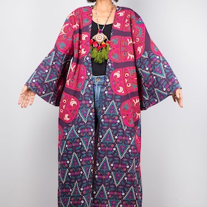Pu&ograve; includere: Un kimono lungo con motivo geometrico viola e rosa. Il kimono ha maniche larghe e una scollatura a V profonda. La modella indossa il kimono sopra una maglietta nera e jeans blu.
