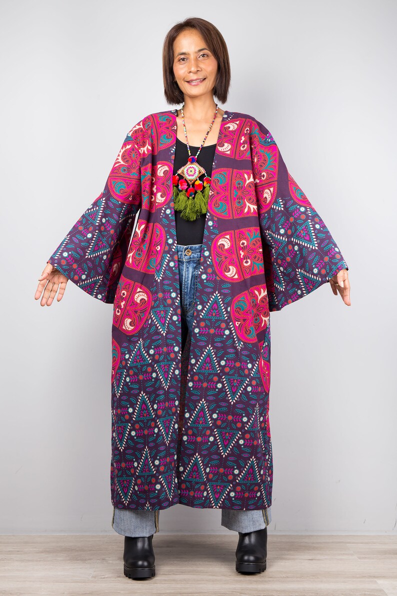 Pu&ograve; includere: Un kimono lungo con motivo geometrico viola e rosa. Il kimono ha maniche larghe e una scollatura a V profonda. La modella indossa il kimono sopra una maglietta nera e jeans blu.