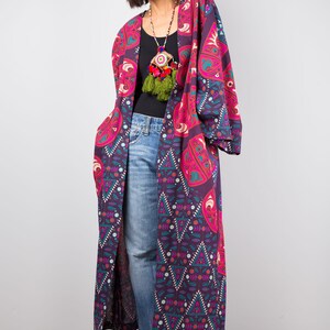 Pu&ograve; includere: Un kimono lungo e fluido con motivi viola e rosa con una collana di nappe colorate. Il kimono ha un design geometrico con accenti floreali.