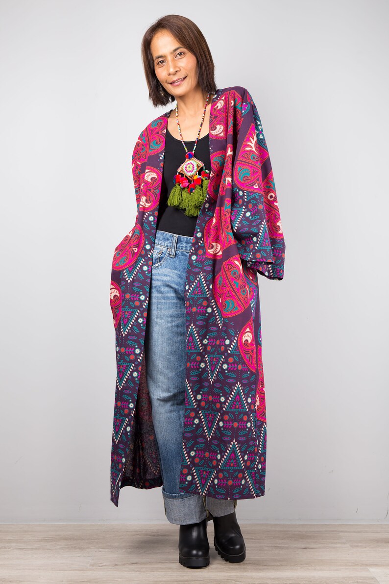 Pu&ograve; includere: Un kimono lungo e fluido con motivi viola e rosa con una collana di nappe colorate. Il kimono ha un design geometrico con accenti floreali.