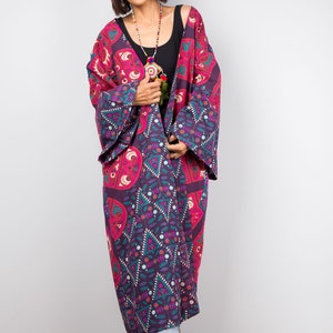 Pu&ograve; includere: Una donna indossa un lungo kimono con motivi viola e rosa con un top nero sotto. Indossa una collana di perline e ha i capelli corti castani.