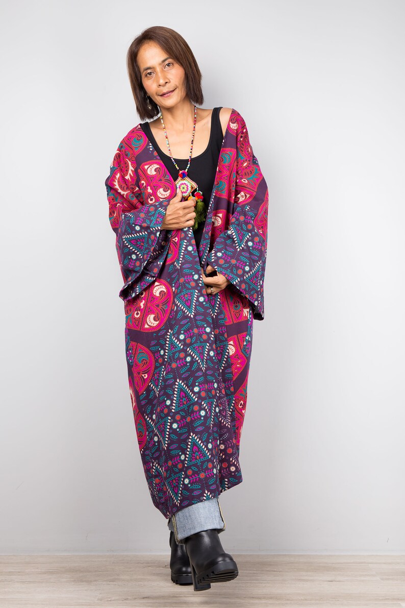 Pu&ograve; includere: Una donna indossa un lungo kimono con motivi viola e rosa con un top nero sotto. Indossa una collana di perline e ha i capelli corti castani.