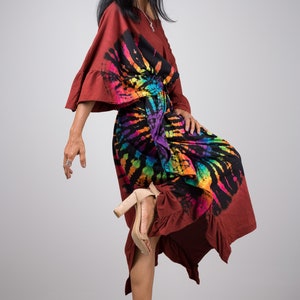 May include: A woman wearing a long, maroon dress with a colorful tie-dye pattern. The dress has a ruffled hem and a loose, flowing fit. She is wearing beige heeled sandals.