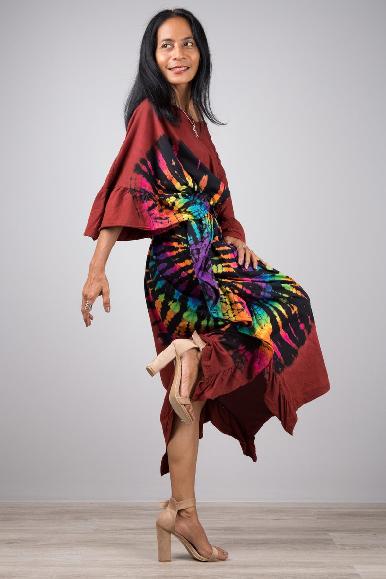 May include: A woman wearing a long, maroon dress with a colorful tie-dye pattern. The dress has a ruffled hem and a loose, flowing fit. She is wearing beige heeled sandals.