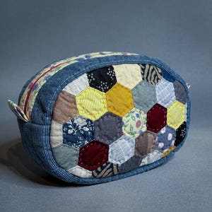 Puede incluir: Un estuche ovalado con cremallera que presenta un exterior de patchwork hecho de varios hexágonos de tela en tonos amarillo, azul, gris, blanco y granate. El estuche está enmarcado por un borde de pana azul texturizada y una pequeña pestaña.