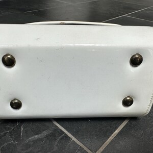 May include: A white patent leather handbag base featuring a rectangular shape with rounded corners and four circular metallic feet attached to the bottom surface to provide stability and protection when the bag is placed on a flat surface.