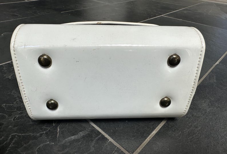 May include: A white patent leather handbag base featuring a rectangular shape with rounded corners and four circular metallic feet attached to the bottom surface to provide stability and protection when the bag is placed on a flat surface.