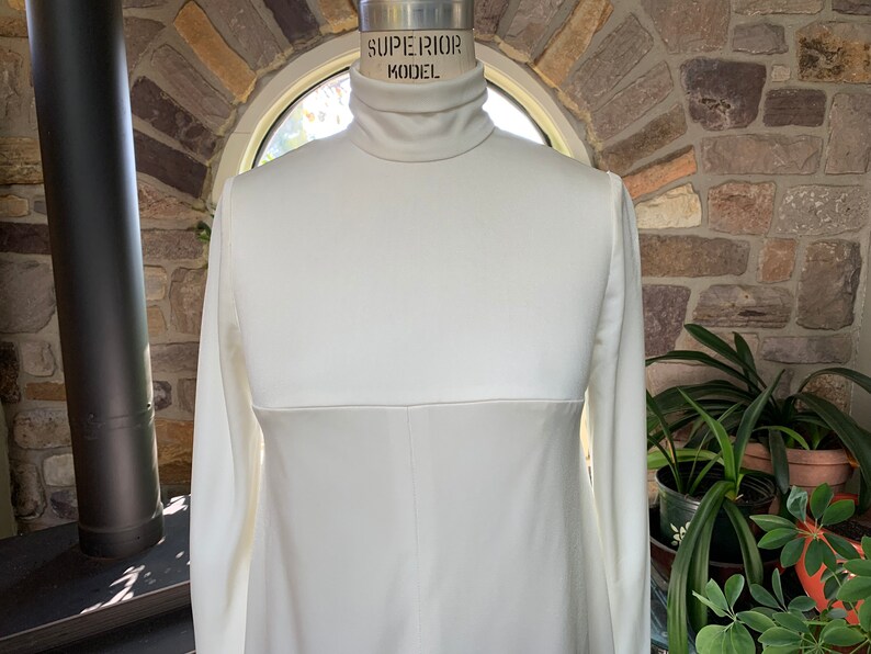 Peut inclure: Une robe cr&egrave;me &agrave; manches longues avec un col roul&eacute;. La robe est pr&eacute;sent&eacute;e sur un mannequin. La robe a une couture sur la section m&eacute;diane. Le mannequin est &eacute;tiquet&eacute; "SUPERIOR MODEL".