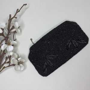 Op de afbeelding: Een zwarte, met kralen bezette clutch met een bladeren ontwerp. De rechthoekige tas heeft een zilveren ketting en is versierd met kleine, donkere kralen. Een takje witte bloemen en een bruine tak staan op de achtergrond.