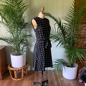 May include: A black and white polka dot dress with a white sash. The dress is on a mannequin in a room with plants.