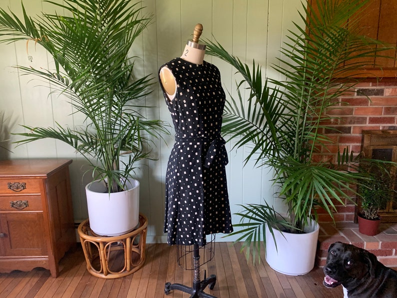 May include: A black and white polka dot dress with a white sash. The dress is on a mannequin in a room with plants.