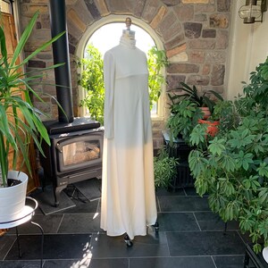 Peut inclure: Une longue robe ivoire &agrave; col montant et manches longues est pr&eacute;sent&eacute;e sur un mannequin. La robe a un design simple et &eacute;l&eacute;gant et une jupe fluide. L'arri&egrave;re-plan comprend un mur en pierre, des plantes et une chemin&eacute;e.