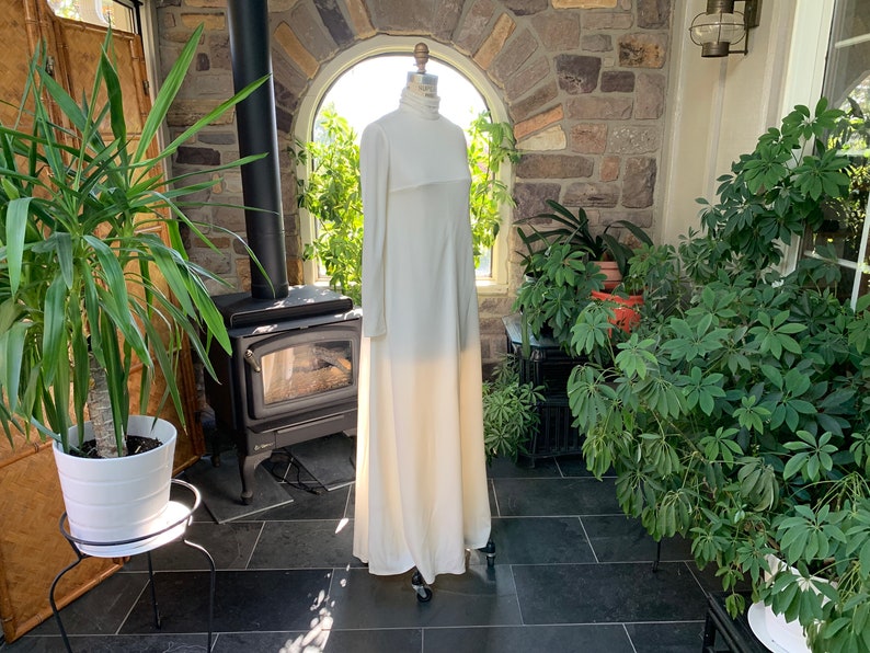 Peut inclure: Une longue robe ivoire &agrave; col montant et manches longues est pr&eacute;sent&eacute;e sur un mannequin. La robe a un design simple et &eacute;l&eacute;gant et une jupe fluide. L'arri&egrave;re-plan comprend un mur en pierre, des plantes et une chemin&eacute;e.