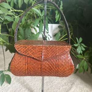 May include: A vintage handbag with a brown snakeskin pattern. The bag features a curved flap closure and a short, dark handle. The bag is displayed against a backdrop of green foliage.