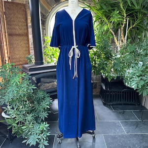 Könnte beinhalten: Ein langes, marineblaues Kleid mit V-Ausschnitt und kurzen Ärmeln. Das Kleid hat weiße Verzierungen am Ausschnitt, an den Ärmeln und eine Taille mit Kordelzug. Das Kleid wird auf einer Schaufensterpuppe präsentiert.