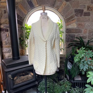 Könnte beinhalten: Eine cremefarbene Langarm-Strickjacke mit breitem Schalkragen, einem strukturierten Strickmuster am gesamten Körper und zwei aufgesetzten Vordertaschen. Das Kleidungsstück wird auf einer Schneiderpuppe präsentiert und hat einen Knopfverschluss.