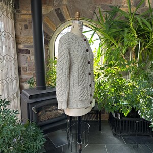 K&ouml;nnte beinhalten: Hellbeige Strickjacke mit Zopfmuster, braunen Kn&ouml;pfen und zwei Vordertaschen. Die Jacke wird auf einer Schaufensterpuppe pr&auml;sentiert. Der Hintergrund zeigt eine Steinmauer, einen schwarzen Ofen und &uuml;ppige gr&uuml;ne Pflanzen.