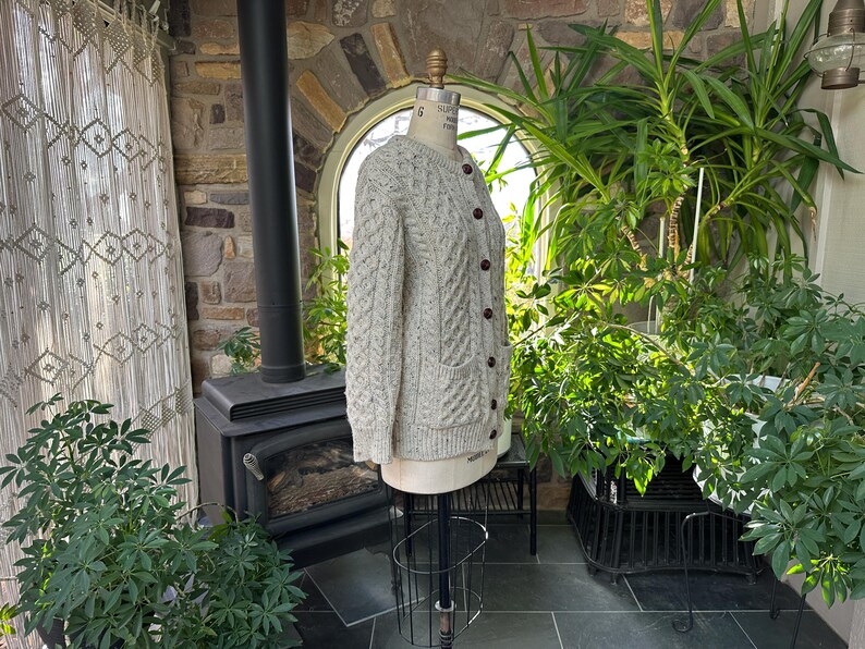 K&ouml;nnte beinhalten: Hellbeige Strickjacke mit Zopfmuster, braunen Kn&ouml;pfen und zwei Vordertaschen. Die Jacke wird auf einer Schaufensterpuppe pr&auml;sentiert. Der Hintergrund zeigt eine Steinmauer, einen schwarzen Ofen und &uuml;ppige gr&uuml;ne Pflanzen.