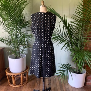May include: A black sleeveless dress with a white geometric pattern. The dress is on a mannequin and is photographed against a green wall.