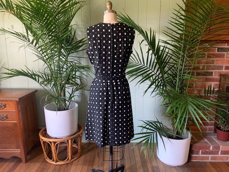 May include: A black sleeveless dress with a white geometric pattern. The dress is on a mannequin and is photographed against a green wall.