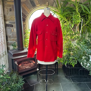 May include: A vibrant red shirt with a collar and two chest pockets, presented on a mannequin. The shirt features button closures and visible stitching. The background includes a fireplace, plants, and a window.