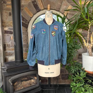 May include: A blue denim bomber jacket featuring a front zipper closure, dark ribbed cuffs, and a ribbed collar. The jacket is decorated with several colorful embroidered patches on the chest and sleeves, including designs of landmarks and nature scenes.