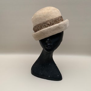 Può includere: Un cappello color crema in stile vintage con una fascia decorativa. Il cappello è realizzato in un materiale morbido e sfocato e presenta una fascia dettagliata color argento attorno alla corona. Il cappello è esposto su una testa di manichino nera.